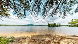 Un lago con orilla arenosa, rodeado de árboles y montañas a lo lejos.