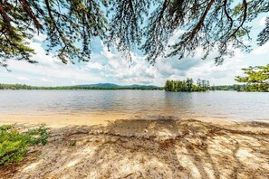 A lake with a sandy shore, surrounded by trees and mountains in the distance. - Homey lakeside property w/ one house, two cottages, mtn. views, & private beach (Ossipee)