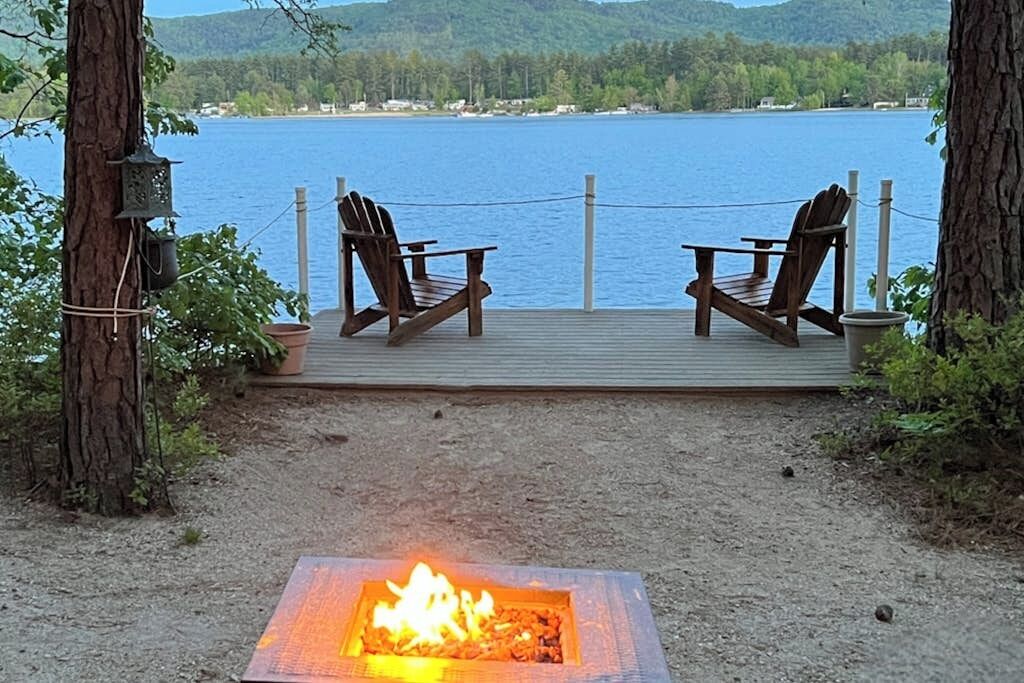 A deck with two chairs overlooking a lake, a fire pit, and a view of the lake and hills.