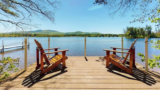 Una terraza de madera con dos sillas Adirondack que dan a un lago tranquilo con montañas al fondo.
