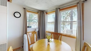 A room with a round wooden table, two windows with curtains, a wall clock, and a radiator. - Tranquil lakefront cottage with private beach, boat slip & fishing kayak access (Ossipee)