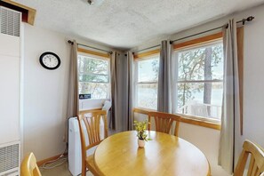 A room with a round wooden table, two windows with curtains, a wall clock, and a radiator. - Tranquil lakefront cottage with private beach, boat slip & fishing kayak access (Ossipee)