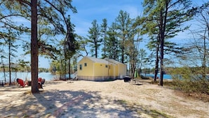 A yellow cabin with a gravel yard, surrounded by trees and a lake. - Tranquil lakefront cottage with private beach, boat slip & fishing kayak access (Ossipee)