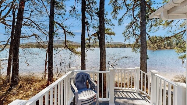 A wooden deck with white railings, overlooking a calm lake with trees and distant hills. - Lakefront home with private beach, access to dock/fishing/kayaks/boat slip (Ossipee)