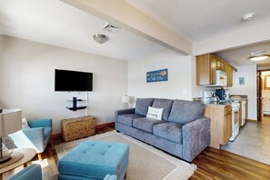 A living room with a grey sofa, a flat-screen TV, and a kitchen area with wooden cabinets. - Lakefront home with private beach, access to dock/fishing/kayaks/boat slip (Ossipee)