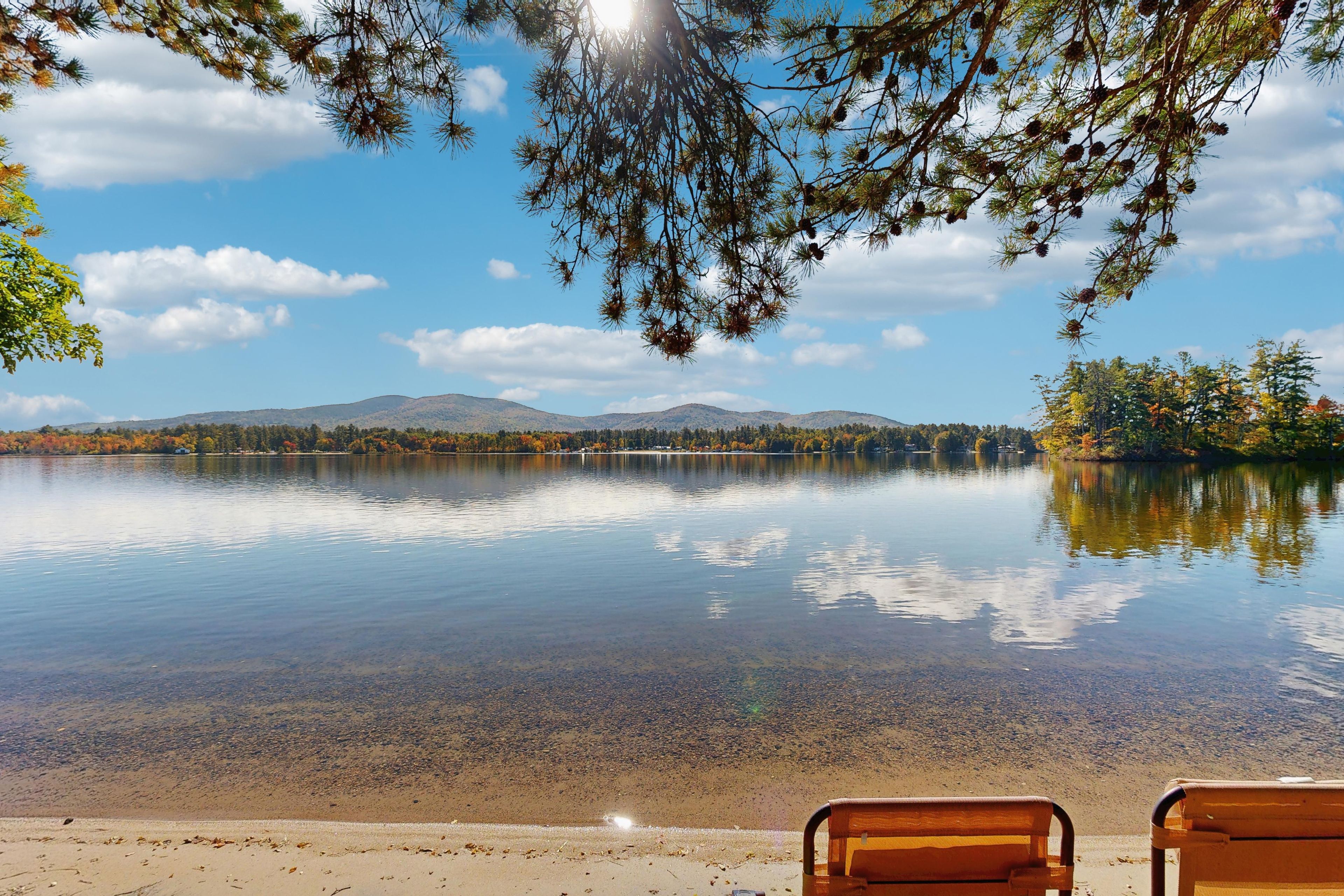 Ruhiger See mit klarem Wasser, das Himmel und umliegende Bäume spiegelt, ein Sandstrand und zwei hölzerne Bänke mit Blick auf das Wasser.