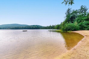 Un lago tranquilo con orilla arenosa, rodeado de un bosque denso y una colina al fondo.