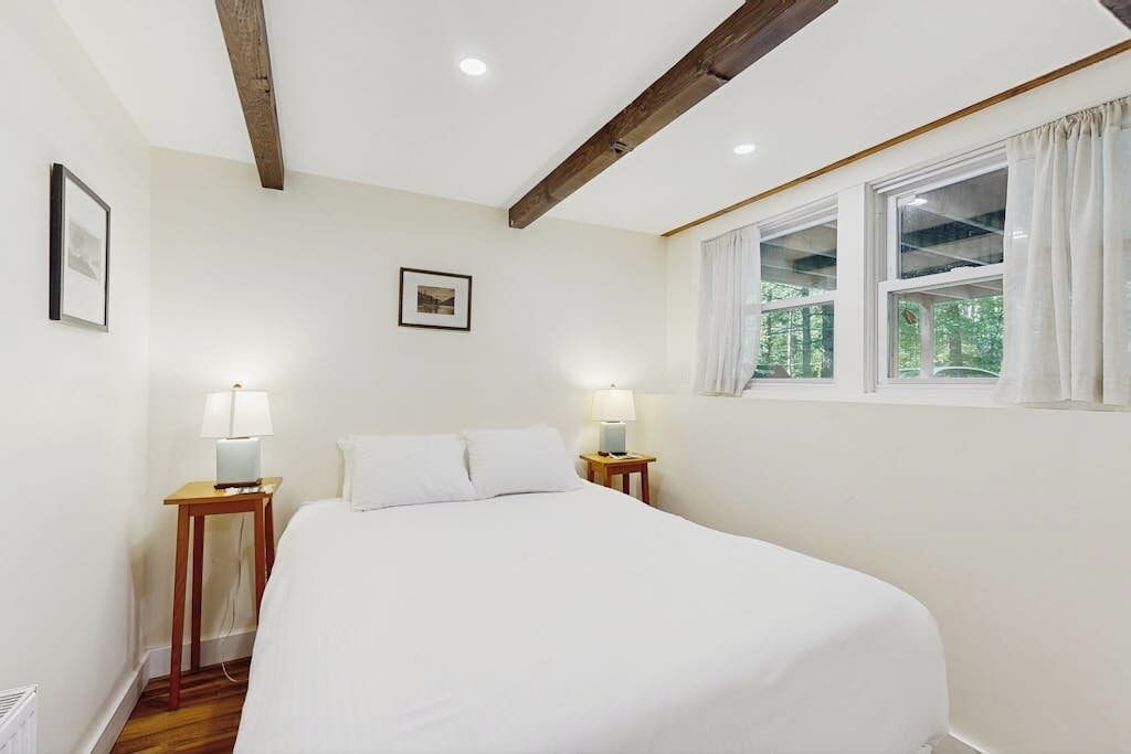 Une chambre à coucher avec un lit, deux tables de chevet avec des lampes, une fenêtre avec des rideaux et un tableau encadré au mur.