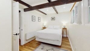 Ein Schlafzimmer mit Bett, Holzbalken, weißen Wänden, gerahmten Bildern, Nachttischlampen und einem Fenster.