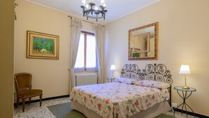 Ein Schlafzimmer mit einem Bett, einem Sessel, einem Fenster mit Vorhängen, einer Heizung, einer Kandelaberleuchte, einem Spiegel und zwei Nachttischen mit Lampen.