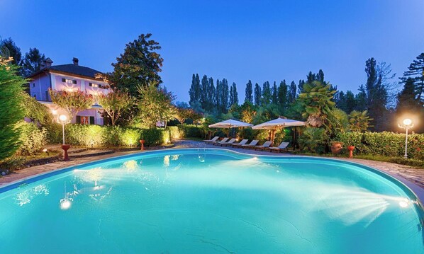 Une piscine entourée d’arbres et d’un bâtiment, avec des chaises longues et des parasols.
