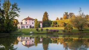 Ein großes, pinkfarbenes Gebäude mit Spiegelung in ruhigem Wasser, umgeben von Bäumen und einer grünen Wiese.