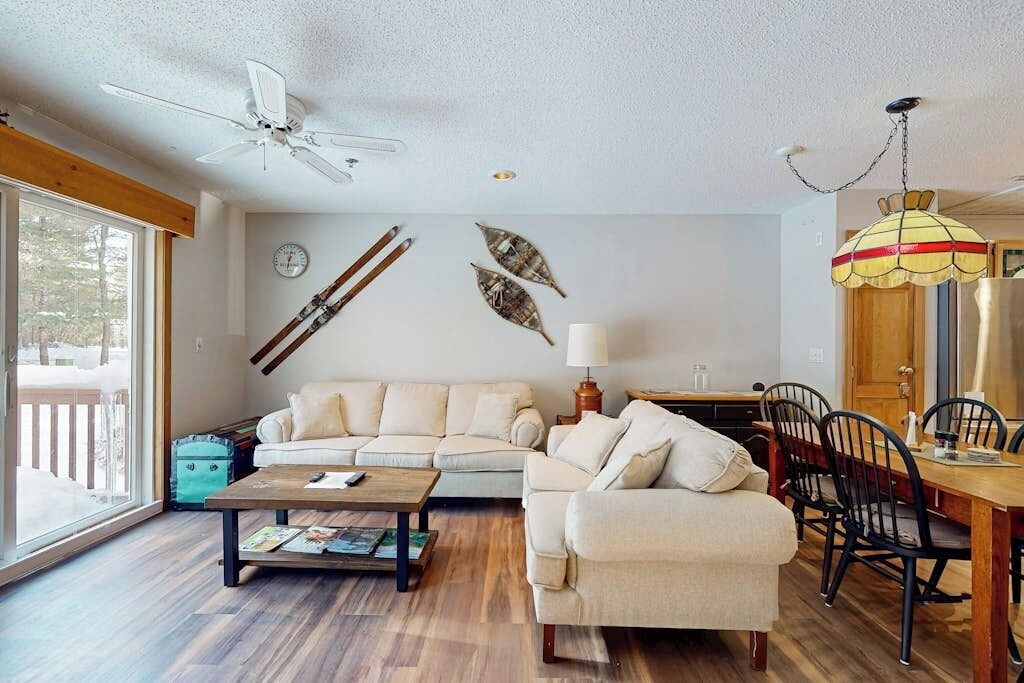 Ein Wohnzimmer mit Sofa, Couchtisch, Esstisch, Stühlen, einem Deckenventilator, einer Lampe und Wanddekoration aus Blättern.