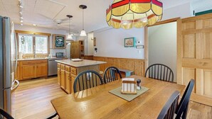 A kitchen with wooden cabinets, a dining table with chairs, a hanging light fixture, and a refrigerator. - Mountain condo: centrally located to shopping, restaurants, skiing, hike & more (North Conway)