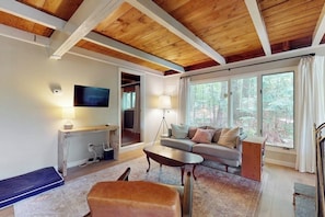 A cozy living room with a wooden ceiling, a sofa, a coffee table, a TV, and a large window with a view of trees. - Country dog friendly lake house w/ access to beaches, tennis & pickleball (Tuftonboro)