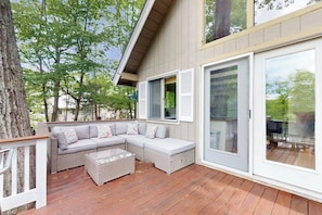 A covered outdoor seating area with a sofa and ottoman, a wooden deck, and a large sliding glass door. - Country dog friendly lake house w/ access to beaches, tennis & pickleball (Tuftonboro)