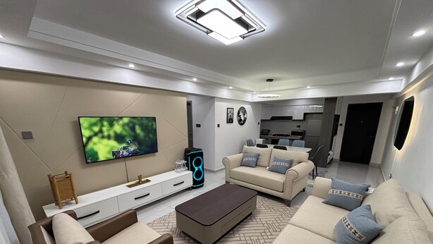 A modern living room with a flat-screen TV, sofas, a coffee table, and a kitchen in the background. - 2-bedroom apartment on 18th floor in adorable Nairobi with fitness room access, (Nairobi)
