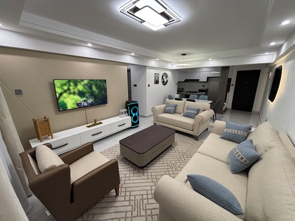 A modern living room with a flat-screen TV, sofas, a coffee table, and a kitchen in the background. - 2-bedroom apartment on 18th floor in adorable Nairobi with fitness room access, (Nairobi)