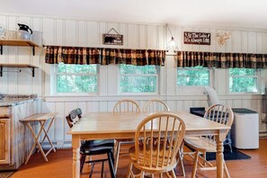 A dining area with a wooden table and chairs, a kitchen area with a stove, and windows with curtains. - Lakefront cottage on Big Pea Porridge Pond w/ private dock, beach, kayaks, canoe (Madison)