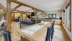 A spacious dining area with a long wooden table, surrounded by chairs, and a kitchen in the background. - Spectacularly updated family mountain lodge w/deck & amazing views (Madison)