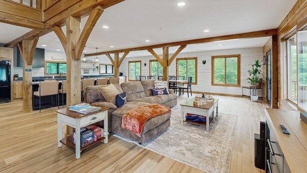 A spacious living area with a high ceiling, wooden beams, a sofa, a coffee table, and a dining area with chairs. - Spectacularly updated family mountain lodge w/deck & amazing views (Madison)