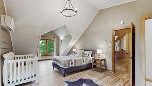 A bedroom with a sloped ceiling, a bed, a white crib, a nightstand, and a view of greenery outside. - Spectacularly updated family mountain lodge w/deck & amazing views (Madison)