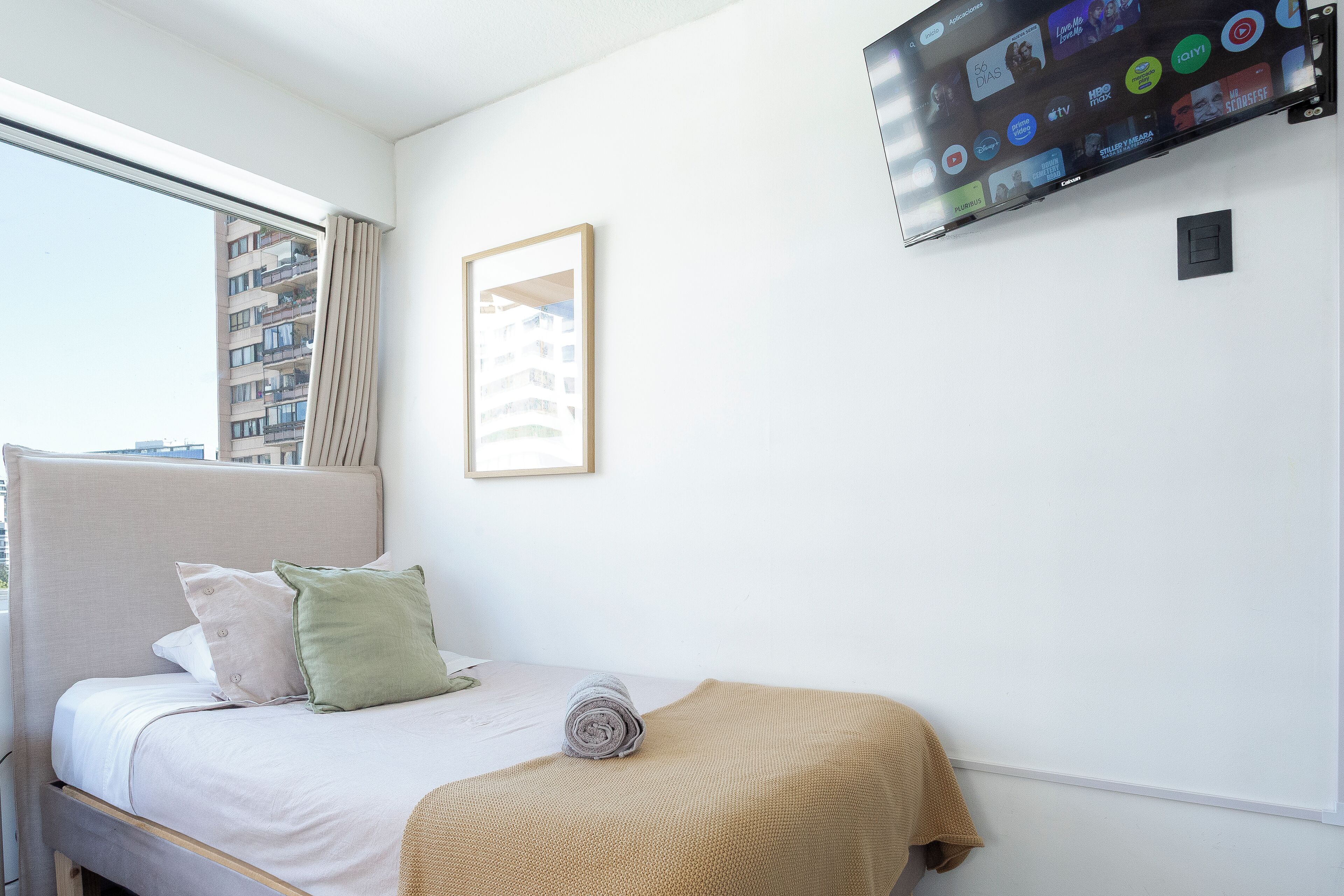 Un dormitorio con cama, televisor montado en la pared, ventana y un cuadro enmarcado.
