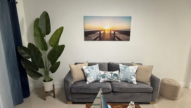 Ein Wohnzimmer mit einer Couch, einem Couchtisch mit einem Modellsegelboot, einer großen Pflanze und einem Sonnenuntergangsbild an der Wand.