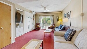 Een hotelkamer met een rood tapijt, een bank, een stoel, een tafel, een televisie, een plafondventilator en een raam met gordijnen.