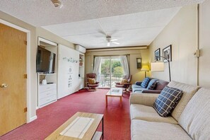 Ein Hotelzimmer mit rotem Teppich, einer Couch, einem Sessel, einem Tisch, einem Fernseher, einem Deckenventilator und einem Fenster mit Vorhängen.