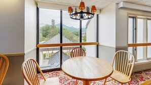 Een kamer met een ronde houten tafel, vier houten stoelen, een kroonluchter, grote ramen en uitzicht op groen en bergen.