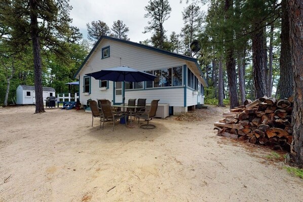 Une cabane dotée d’un patio couvert, d’une table à manger avec des chaises et d’une pile de bois de chauffage.