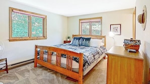 A bedroom with a wooden bed, two windows, a bedside table, a lamp, and a wall-mounted hat.