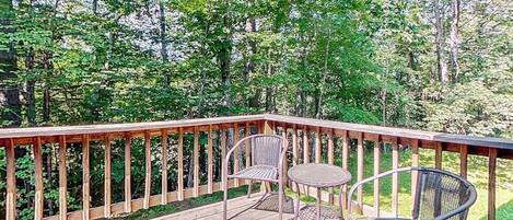 A wooden deck with metal chairs and a small table, surrounded by dense green foliage.