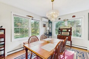 A dining room with a wooden table, chairs, a chandelier, and a view of greenery outside. - Private spacious mountain view w/ beach rights, hot tub, spacious deck & firepit (Ossipee)