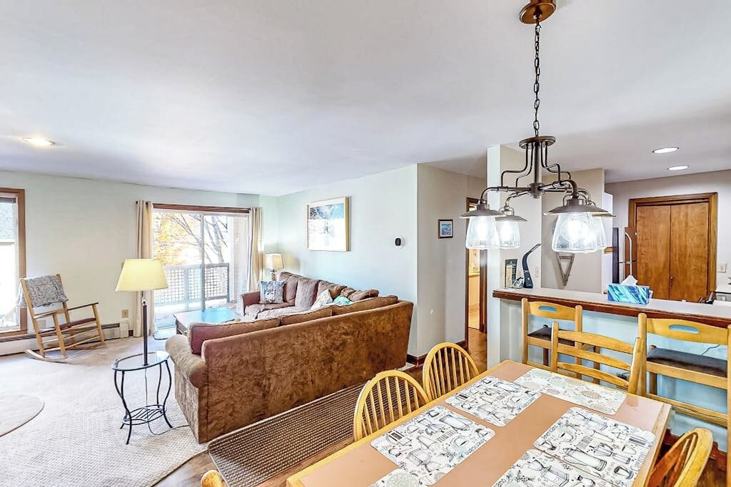 Un salon avec un canapé, une table à manger et des chaises, un lustre et un coin cuisine avec un meuble en bois.