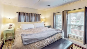 Una habitación de hotel con una cama grande, lámparas de noche, un banco y una ventana con cortinas.