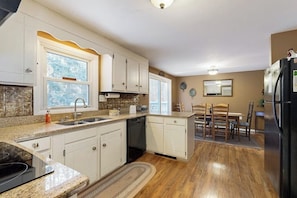 A kitchen with white cabinets, a granite countertop, a black refrigerator, and a dining area with wooden chairs. - Mountain home near shops, hiking, skiing, restaurants & brewery (North Conway)