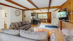 A living room with a sofa, a television, and a kitchen in the background. - Intown home w/ views, private deck & firepit; close to  Mt. Cranmore (North Conway)