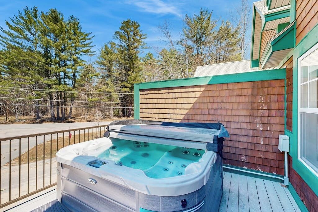 A hot tub on a wooden deck with a metal railing, surrounded by trees and a building with green trim.