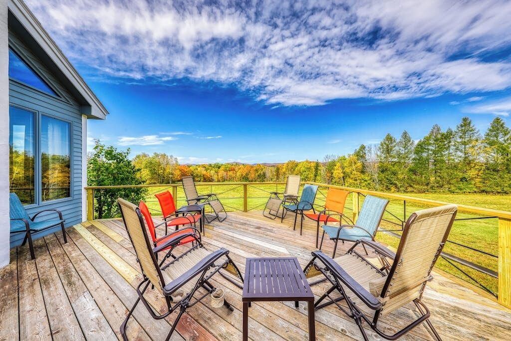 Une terrasse en bois avec des meubles d’extérieur, une vue sur un champ verdoyant et un bâtiment bleu.