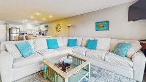 Un salón con un sofá seccional, una mesa de centro, una cocina al fondo y un reloj de pared.