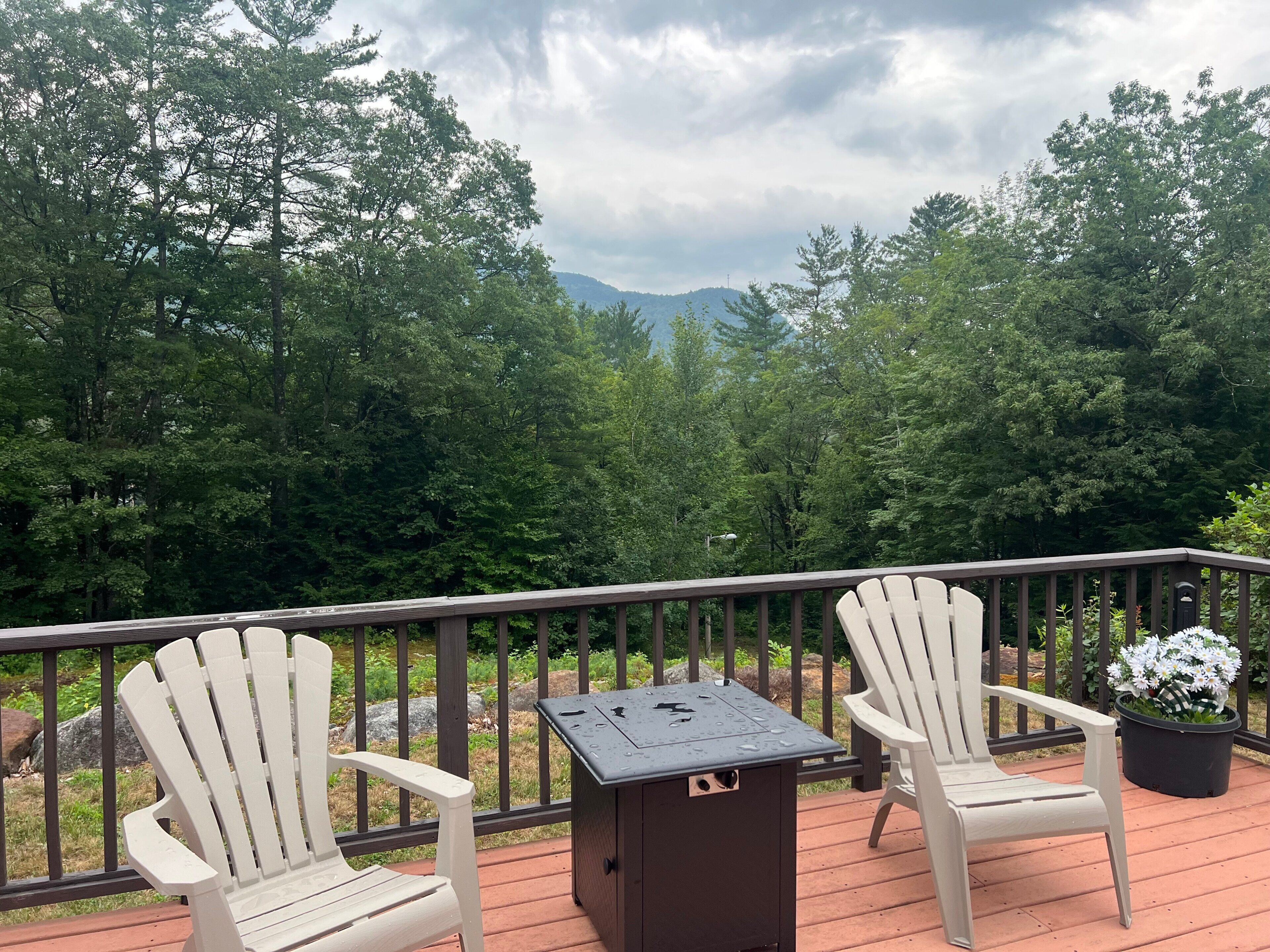 Um terraço com duas cadeiras Adirondack brancas e uma churrasqueira, com vista para uma área arborizada e montanhas ao longe.