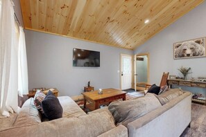 Ett mysigt vardagsrum med ett träpanelat tak, en tv, en soffa, ett soffbord och en dörr som leder till ett annat rum.