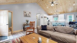 A spacious living area with a high ceiling, a wooden ceiling, a sofa, a coffee table, a chair, a kitchen in the background, and a door leading to another room.