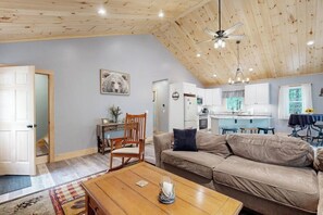 Ett rymligt vardagsrum med högt i tak, ett träpanelat tak, en soffa, ett soffbord, en fåtölj, ett kök i bakgrunden och en dörr som leder till ett annat rum.