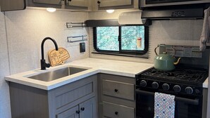 Cozinha compacta de motorhome, com pia, fogão, micro-ondas e janela com vista.