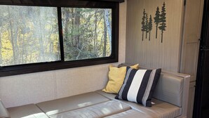 Interior moderno de uma caravana, com sofá-cama, janela com vista para uma floresta e armário fixado na parede.