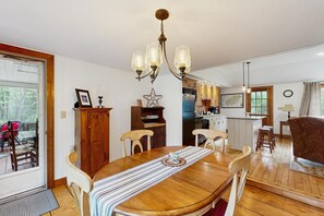 Uma sala de jantar com mesa de madeira, cadeiras, um candelabro e vista para a cozinha e a sala de estar.