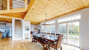 Un vaste espace dédié à la salle à manger, avec une table et des chaises en bois, un escalier menant à un niveau supérieur et de grandes fenêtres offrant une vue sur l’extérieur.